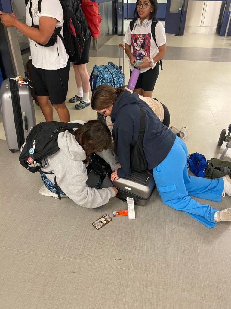 three girls sit on a suitcase to try to close it